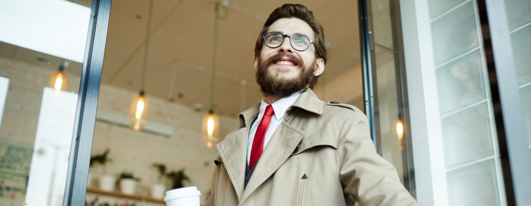 Smiling bearded businessman in trench coat entering glass door with coffee and briefcase