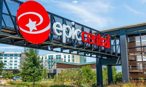 A large red and white EpicCentral sign mounted on a black metal pedestrian bridge overlooking a landscaped walkway in Grand Prairie.